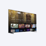 نمای کمی زاویه‌دار از یک تلویزیون صفحه تخت که رابط کاربری Google TV را نمایش می‌دهد. صفحه نمایش "Lord of the Rings: The Rings of Power" را به عنوان عنوان اصلی نشان می‌دهد، و چندین برنامه سرویس پخش دیگر مانند YouTube، Prime Video و Disney+ در پایین قابل مشاهده هستند. تلویزیون توسط دو پایه تیره رنگ خاکستری پشتیبانی می‌شود.